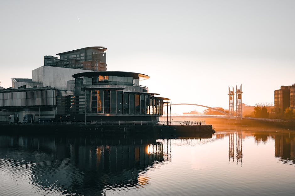 A modern, multi-story building with large glass windows and a rounded, layered architectural design is situated on the edge of a calm waterway, with its reflection visible on the surface. Adjacent to the building, a pathway or loading area features a stack of cardboard boxes wrapped in plastic and fabric moving blankets, along with a trolley and various packing materials. A person is seen lifting or carrying a cardboard box towards a parked van or moving vehicle positioned nearby for a home relocation or furniture transport process. The area is illuminated by warm sunlight during what appears to be late afternoon or early evening, with additional structures and industrial cranes visible in the background, suggesting an urban environment. This scene exemplifies the loading process involved in house removals and packing services, indicating a professional move facilitated by [COMPANY_NAME] as part of their removals offering in the Surrey Quays area.