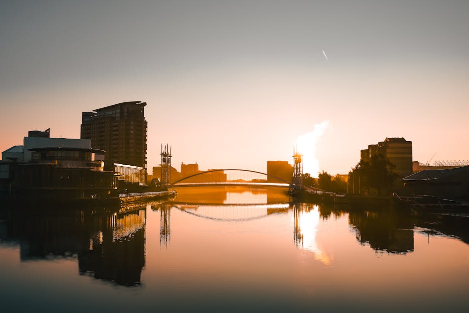 A panoramic view of the Surrey Quays area at sunset, featuring modern high-rise buildings on the left side and smaller structures along the water's edge. In the foreground, calm river water reflects the silhouettes of the buildings and the sky's warm tones. A bridge spans across the river in the background, with a thin trail of steam or smoke rising from a chimney or industrial site on the right. The scene is illuminated by natural sunset light, casting a golden hue over the scene, and the environment appears tranquil, with no visible movement or activity. This scenic urban landscape showcases the mixture of residential and commercial architecture typical of Surrey Quays, often involved in home relocation and furniture transport logistics, as handled by companies like Man with Van Surrey Quays during comprehensive packing and loading processes.