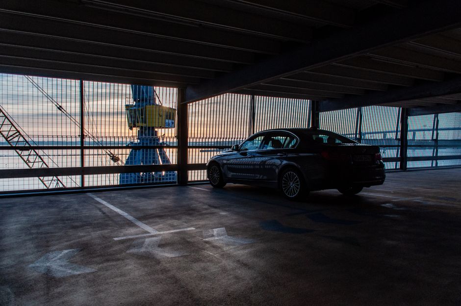 A dark-colored sedan is parked in an empty, covered parking space within a multi-storey car park overlooking a construction crane and the surrounding area. The parking space shows painted lines and numerical markings on the concrete floor, with some areas reflecting ambient light. The metal fencing along the open side of the parking area reveals the exterior environment, including the crane and a sunset sky with clouds. The interior of the parking garage features a low ceiling with beams and a subdued lighting atmosphere. This setting illustrates the logistics involved in home relocation, with a focus on the importance of proper parking and loading arrangements for furniture transport during a house move facilitated by Man with Van Surrey Quays, especially when coordinating in tight or restricted loading bay areas.