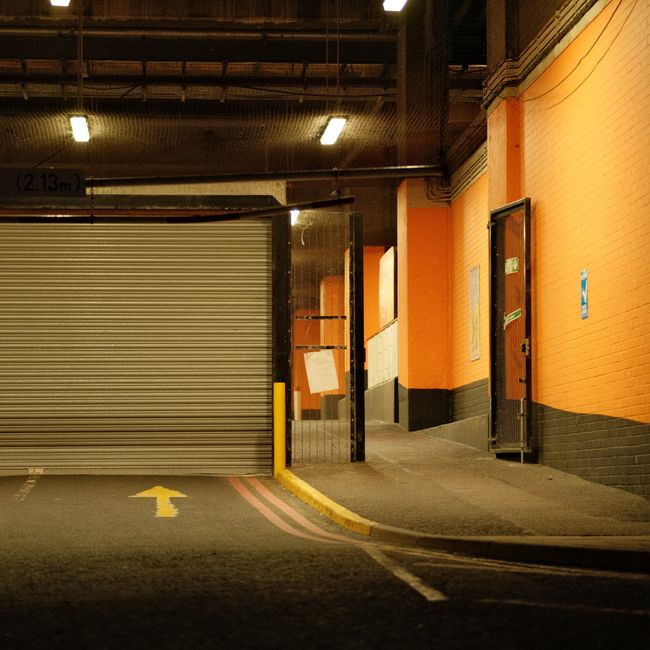 Inside a building's loading bay, a partially open metal roller shutter door reveals a narrow passage leading to a storage or service area. The surrounding walls are painted orange with black protective skirting at the base. Adjacent to the entrance, a small door with a vertical window and a handle stands open, revealing a glimpse of the interior. The concrete pavement outside features a yellow arrow painted on the ground, indicating directional flow for vehicle or pedestrian movement, and a yellow curb edge guides the loading process. Overhead lighting illuminates the space with bright, artificial light, casting shadows on the floor. The image captures a clean, organized environment suitable for home relocation or furniture transport, with a focus on efficient loading and unloading in a commercial or residential building managed by Man with Van Surrey Quays. This setting supports professional removals and packing activities essential to effective house removals and loading bay access, aligning with the topic of avoiding fines through proper loading bay usage during Surrey Quays moves.