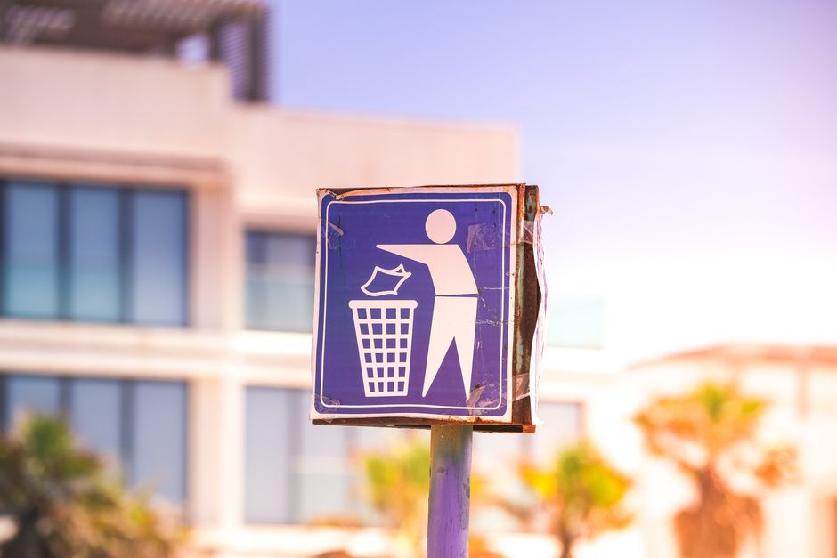 A close-up of a weathered blue city waste disposal sign mounted on a metal pole outside a residential building. The sign features a white pictogram of a person discarding a piece of paper into a trash bin, indicating proper waste disposal. In the background, blurred modern apartment windows and part of a building facade are visible, with natural daylight illuminating the scene. The image captures the outdoor environment near a home, relevant to moving and packing processes, and emphasizes responsible waste management during home relocation with occasional mentions of [COMPANY_NAME] and their removals services, including furniture transport and logistics in Southwark.