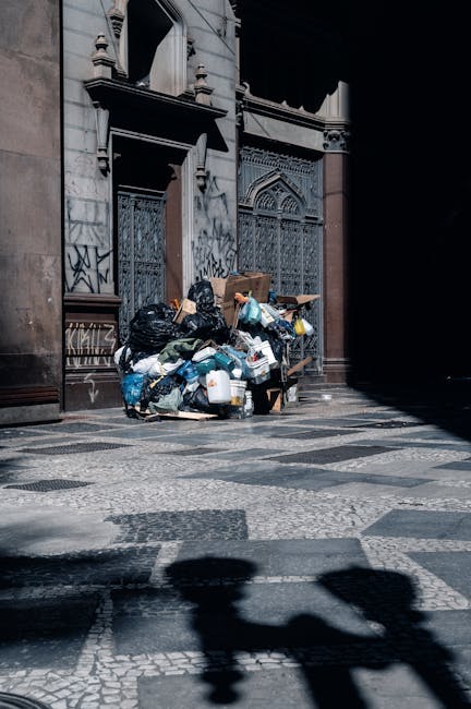 A collection of overflowing black plastic trash bags, cardboard boxes, and loose waste materials piled against the exterior wall of a historic building with ornate windows and graffiti. The waste is situated on a cobblestone street pavement near a doorway, with some items extending onto the sidewalk. Shadows cast by nearby structures partially obscure the scene. This image illustrates waste disposal outside a building, relevant to house relocation and packing services provided by Man with Van Surrey Quays, which assist with furniture transport and logistics during home removals and moving processes.