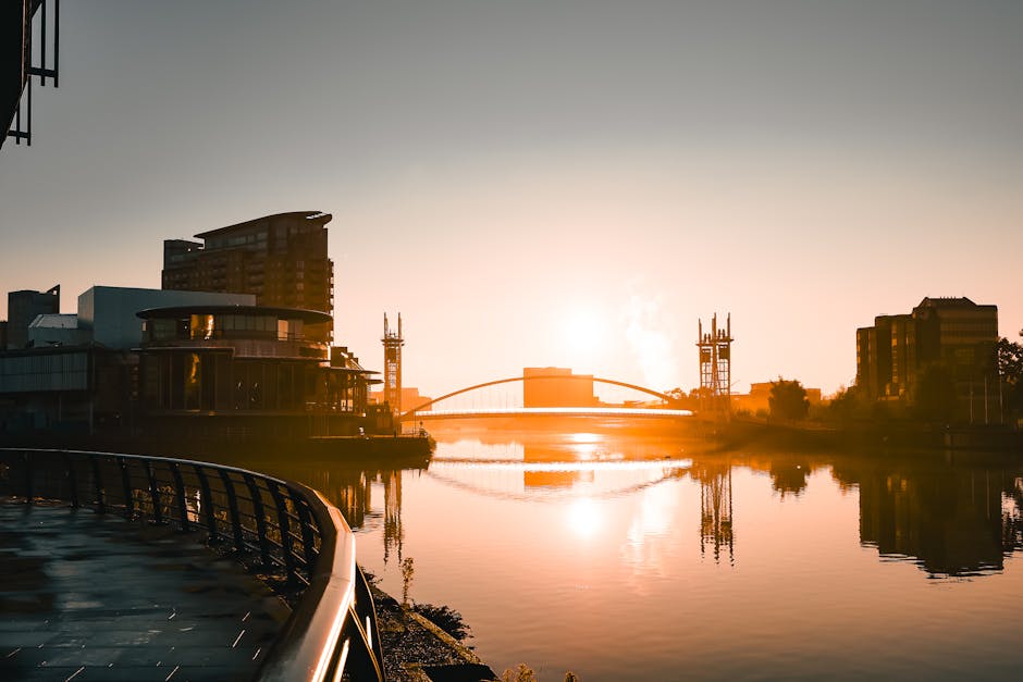 A panoramic view of Surrey Quays at sunrise featuring a calm river with reflections of surrounding modern buildings, including a multi-storey structure with glass windows and a curved roof, and tall communication towers in the background. The scene is illuminated by the rising sun, casting a warm glow over the water and the cityscape. On the left, part of a metal railing along the riverbank is visible, indicating a walkway or promenade. The outdoor environment suggests a peaceful setting suitable for a home relocation or furniture transport process, with the natural light highlighting the area where [COMPANY_NAME] might assist with loading or unloading residential furniture and packed boxes in preparation for moving services, emphasizing the logistics involved in packing and moving activities near the Dockside Moves at Surrey Quays Shopping Centre.
