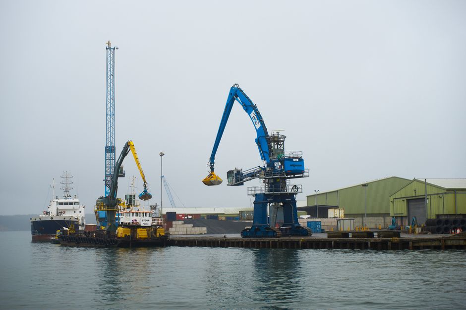 A port scene showing two large cranes equipped with loading arms, one yellow and one blue, positioned on a concrete dock with containers and equipment around. The cranes are actively engaged in loadin