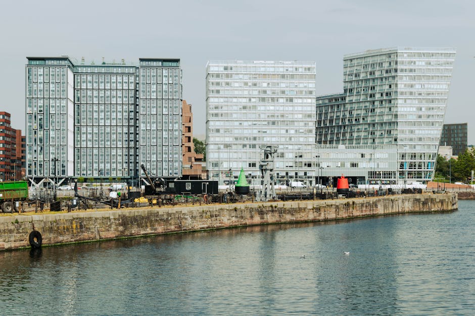 A row of modern high-rise buildings along the waterfront in Greenwich, Surrey Quays, with large glass facades and irregular geometric designs. In the foreground, there is a concrete quay wall with a black rubber tire fender attached, and a narrow strip of water with a few small buoys floating on the surface. Along the pathway on the quay, several black and green shipping containers, mobile cranes, and construction equipment are visible, indicating ongoing development or loading activities related to home relocation or furniture transport. The buildings are illuminated by natural daylight, with a clear sky overhead. This scene illustrates the urban environment and logistic aspects associated with moving services, such as loading and transportation, in the Surrey Quays area, which Man with Van Surrey Quays may support with removals and moving logistics.