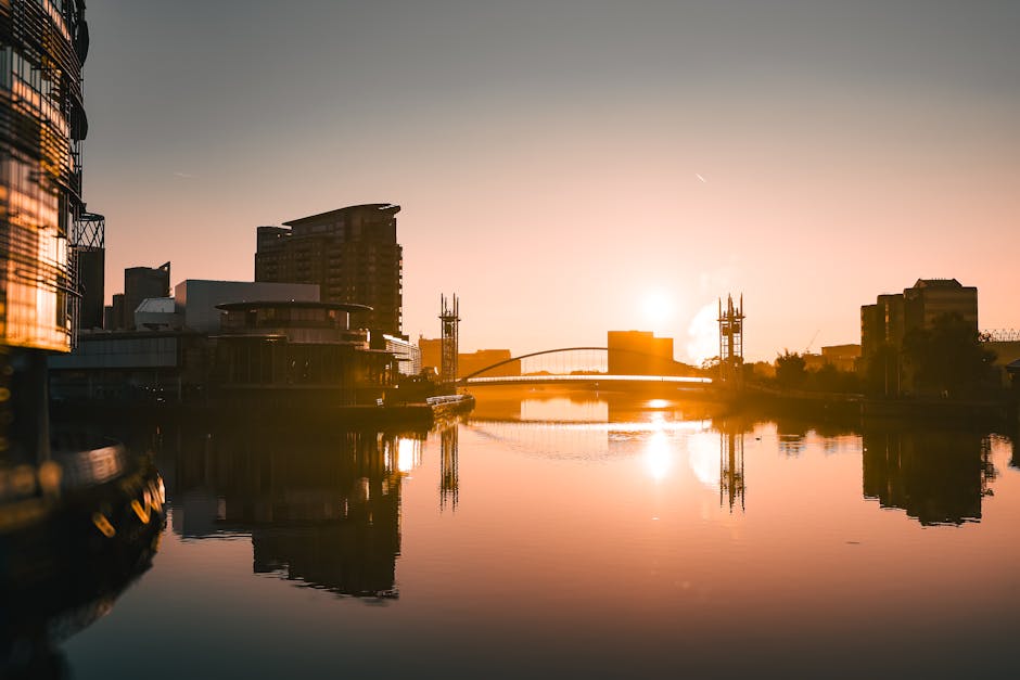 A scenic view of the Greenland Dock area at sunset, featuring modern commercial and residential buildings along the water's edge, with some structures reflecting on the calm river surface. In the background, a bridge with two towers spans across the river, and a few boats are docked near the waterfront. The sky is illuminated by the setting sun, casting warm orange and pink hues over the scene. The image captures the urban landscape and waterfront environment, which may be relevant to house removals and home relocation services provided by Man with Van Surrey Quays that include furniture transport and loading processes involved in moving logistics.