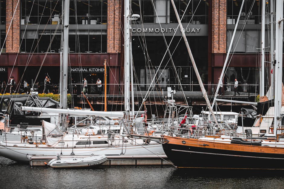 A row of modern high-rise buildings along the waterfront in Greenwich, Surrey Quays, with large glass facades and irregular geometric designs. In the foreground, there is a concrete quay wall with a black rubber tire fender attached, and a narrow strip of water with a few small buoys floating on the surface. Along the pathway on the quay, several black and green shipping containers, mobile cranes, and construction equipment are visible, indicating ongoing development or loading activities related to home relocation or furniture transport. The buildings are illuminated by natural daylight, with a clear sky overhead. This scene illustrates the urban environment and logistic aspects associated with moving services, such as loading and transportation, in the Surrey Quays area, which Man with Van Surrey Quays may support with removals and moving logistics.