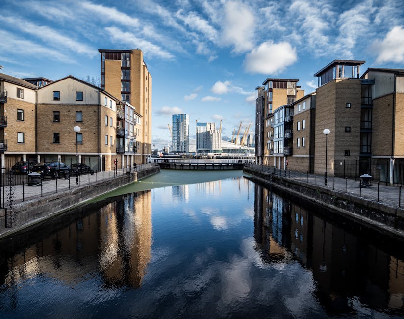 A view of a canal lined with modern residential buildings constructed from beige brick and dark metal accents, with large windows and small balconies. The canal water reflects the buildings and a partly cloudy blue sky. On the left side, parked cars are visible along a paved walkway with black metal railings, and on the right, similar residential structures are present. In the background, a bridge spans the canal, and beyond are high-rise office or apartment buildings with glass facades, indicative of a city urban environment. The scene features natural daylight and clean, maintained surroundings, suggesting an active residential or commercial area within the Surrey Quays locality. This setting is relevant to house removals and furniture transport, as it shows a typical urban canal-side environment where packing and loading processes by [COMPANY_NAME] could take place during a home relocation or moving service, with visible equipment and organized spaces facilitating furniture and box loading activities.
