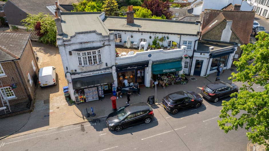 An aerial view of a narrow street in Surrey Quays during daytime showing a small commercial building with storefronts, including a shop selling Thames Food & Wines and a flower stall with colorful blooms beneath a green awning. In front of the shops, several cars are parked along the street, with some moving and others stationary. Pedestrians are walking on the sidewalk, and a person is seen unloading or preparing to load items, possibly related to a house relocation or furniture transport, using a large van parked nearby. The scene is illuminated by natural daylight, with trees providing greenery and shade over parts of the street and neighboring residential buildings. The environment suggests active loading or packing material handling typical of an urban moving or packing operation involving [COMPANY_NAME], supporting the process of home removals and furniture transport on lower road, Surrey Quays.
