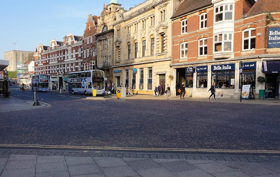 A street scene on Lower Road in Surrey Quays, featuring traditional brick and stone buildings with ornate architectural details, housing various commercial establishments including a pizza restaurant called Bella Italia with its blue sign and awning. The street has a wide paved area and a dedicated bus lane with two double-decker buses parked alongside the kerb. Pedestrians, some carrying bags, are walking on the pavement and crossing the street during daylight with clear weather. In the foreground, there are cardboard boxes, wrapped furniture, and plastic packing materials placed on the pavement, indicating a house removal or relocation process. A vehicle, possibly a van used by Man with Van Surrey Quays, is nearby with open doors, and a hand truck or trolley may be visible, supporting furniture transport and loading activities. The scene reflects the logistical aspects of home relocation, including packing, moving, and street-level logistics typical of a professional removals service in an urban environment.
