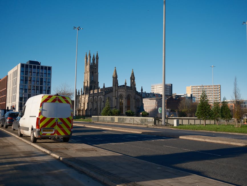 A white commercial van with yellow and red chevron safety markings on its rear doors is parked on the side of a city street, adjacent to a sidewalk with a curb. The van appears to be involved in home relocation or furniture transport, positioned in a loading area near a church with pointed Gothic-style spires, set against a clear blue sky. Around the van, there are other parked vehicles along the road, which has multiple lanes separated by a paved median. Streetlights and modern office buildings are visible in the background, along with trees and a small grassy area. The scene captures a moment during packing and moving activities, possibly part of a larger home removal process, supported by [COMPANY_NAME], which offers professional removals services. The environment is well-lit with natural daylight, emphasizing the urban setting and the logistics involved in a house move or furniture transport along this route.