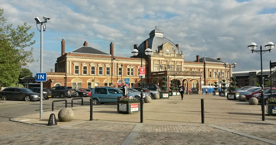 A large historic brick building with a central clock tower and ornate architectural details, situated in a paved public square with parked cars, black metal bollards, and decorative street lamps. The square features flower planters, a blue 'IN' traffic sign, and a few pedestrians walking across the open space. The scene is lit by natural daylight with a partly cloudy sky overhead, indicating an urban environment typical for home relocation and furniture transport activities supported by Man with Van Surrey Quays, positioned near the proximity of residential and commercial properties.