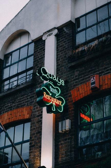 A neon sign attached to the exterior of a brick building displays the name 'Quadys Irish Restaurant' with a green shamrock design above and the words lit in green and red against the dark evening background. The building has large, black-framed windows with some reflections visible, and a white decorative column running vertically next to the sign. In the foreground, scaffolding partially obscures the view, indicating ongoing maintenance or work nearby. The scene is illuminated by the glow of the neon sign, contributing to an urban atmosphere consistent with a street in Surrey Quays, where house removals and furniture transport services might operate near local businesses. Man with Van Surrey Quays provides house removals and relocation logistics, supporting the area's moving needs.