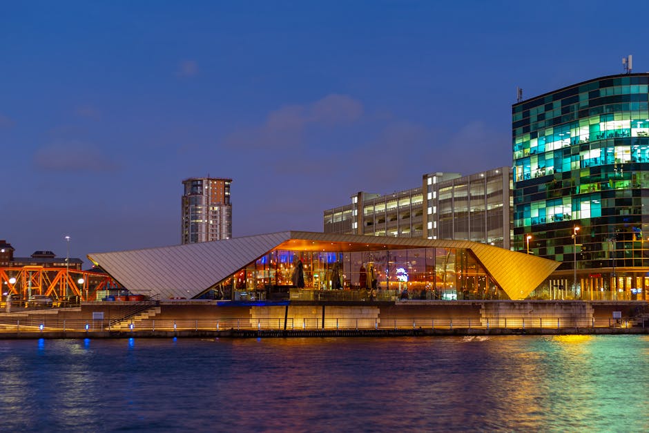 A modern waterfront building in Surrey Quays during the evening, with its glass facade illuminated by interior lighting and reflections on the water. The structure features a distinctive angled, metallic roof extending over a glass-walled interior, with visible tables and chairs inside. Behind it, there are multi-storey office or apartment buildings with lit windows, including a higher-rise with a grid of illuminated panes and a circular building with a curved glass exterior. The scene captures a typical home relocation or furniture transport environment, focusing on the exterior setting of a key location for removals services. The surrounding area is calm, with gentle water reflecting the city lights and a clear evening sky overhead. Man with Van Surrey Quays might utilize such locations for logistical purposes during packing and loading processes for house removals, illustrating the urban environment involved in moving and transport activities.