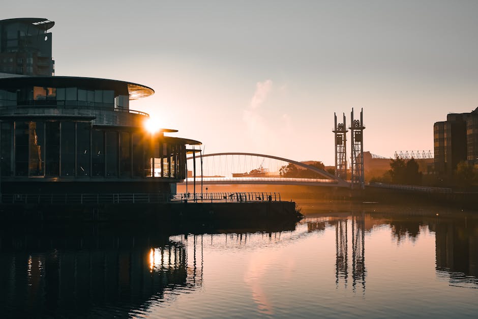 A panoramic view of the Surrey Quays area during sunset, showing modern buildings including a tall multi-storey structure on the left and other commercial or residential buildings on the right. In the foreground, a curved metal railing runs along the edge of a waterfront pathway. The river reflects the warm orange and yellow hues of the setting sun, with a small boat moored nearby. The scene includes a bridge with towers and an arched design spanning the river in the distance, as well as various telecommunications infrastructure, such as antennas, on the buildings. The overall atmosphere is calm, with soft natural lighting highlighting the urban riverside environment, suitable for illustrating home relocation or furniture transport processes involved in house removals in Surrey Quays.